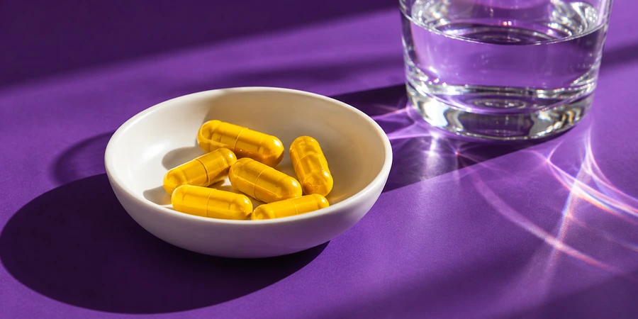 Gélules jaunes sans marque dans une petite coupelle à côté d’un verre d’eau sur un fond violet